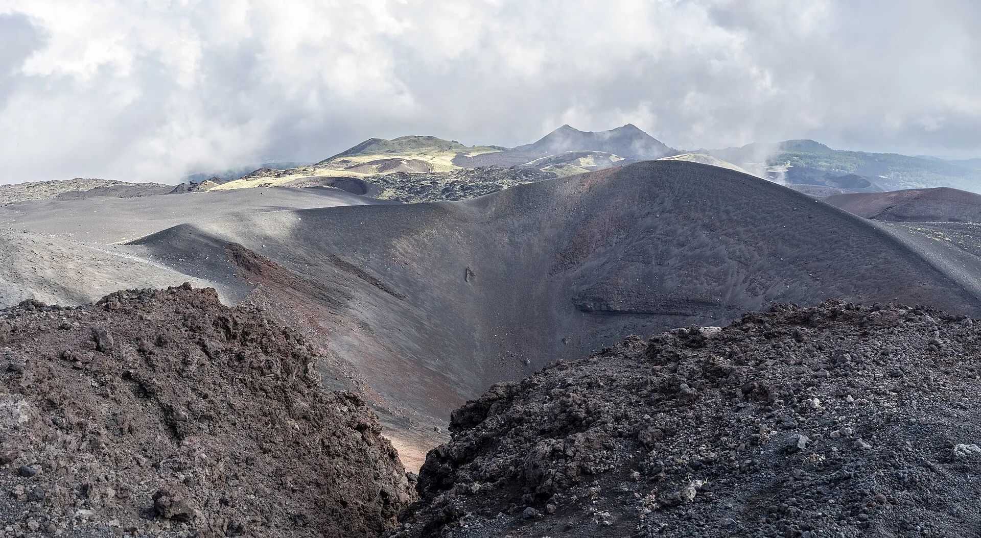 The rim of a summit crater on Etna: dark cinder ramparts, steaming craters and clouds behind