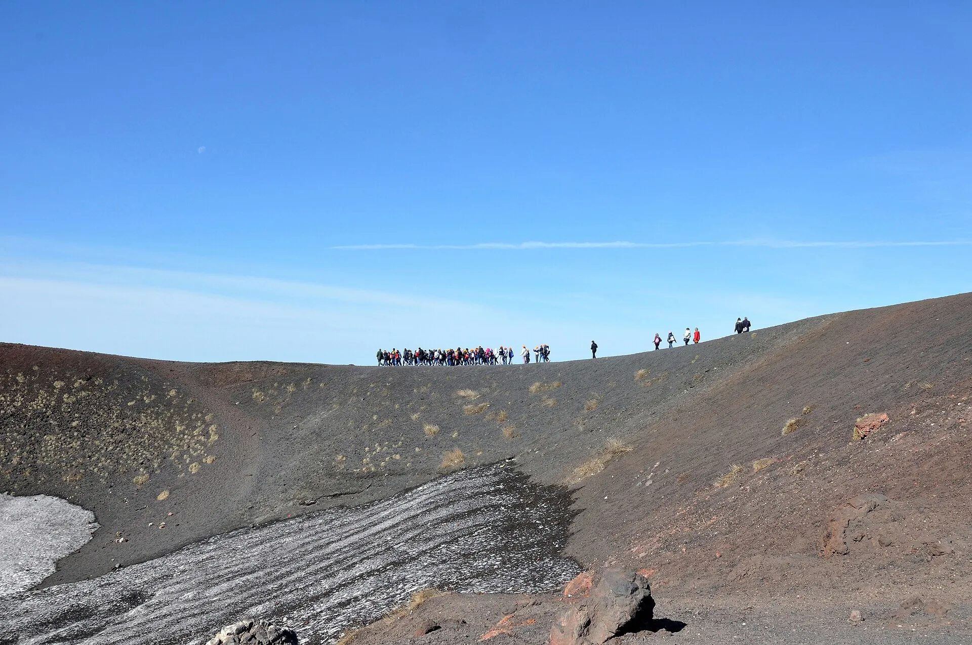 A line of hikers walking along the ridge of the Crateri Silvestri on Etna, black cinder, deep blue sky
