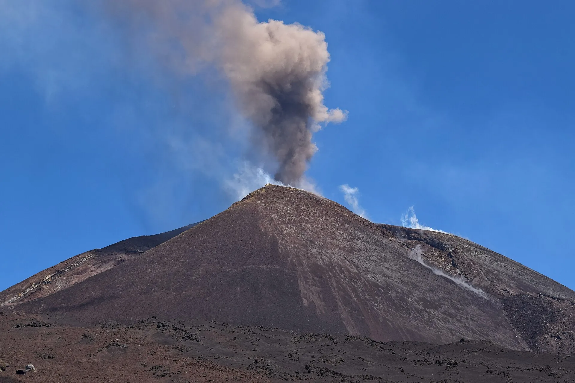 Etna's Southeast Crater sending a grey column of ash into the blue sky, the flank of dark cinder