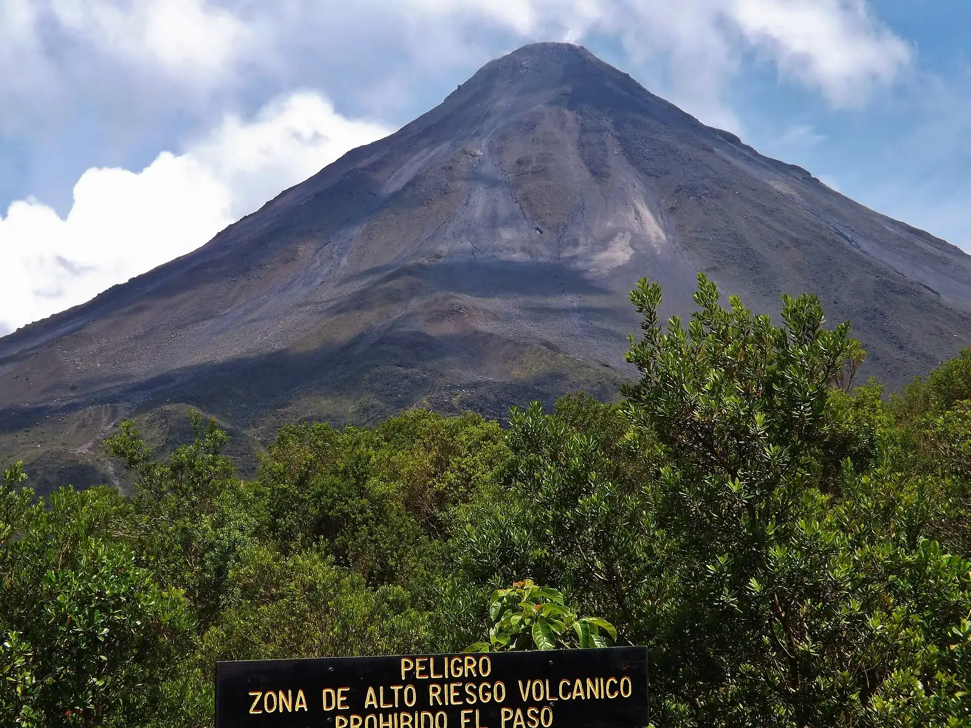 The gray cone of Arenal Volcano above scrubby forest, a warning sign in the foreground reading: Peligro, Zona de alto riesgo volcánico, prohibido el paso