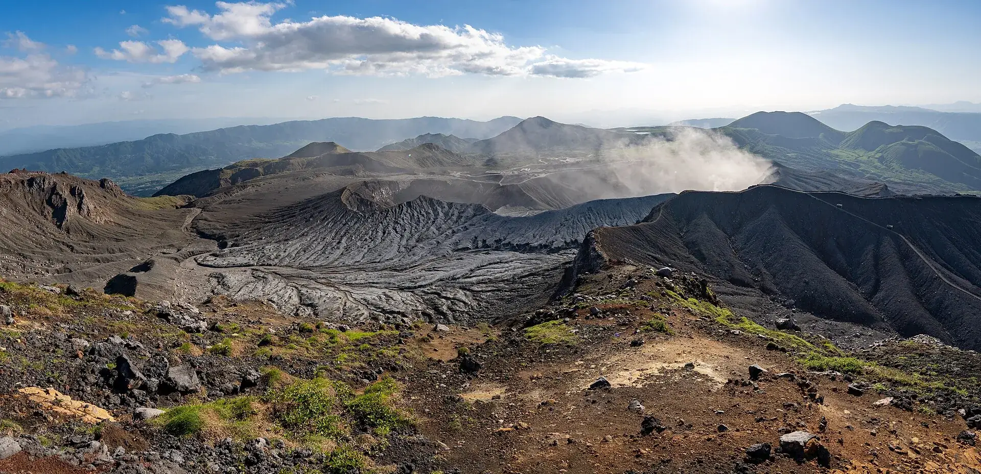 Panorama vom Kraterrand des Aso über graue Aschefelder und die zentralen Gipfel, rechts eine Dampffahne