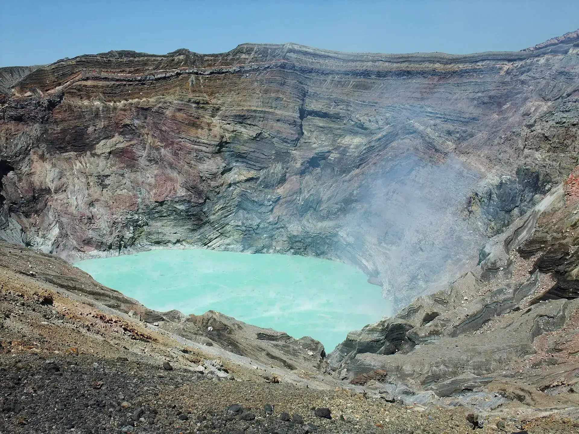 Der türkisgrüne Kratersee im Nakadake-Krater des Aso, umgeben von rot-grau geschichteten Kraterwänden mit aufsteigendem Dampf