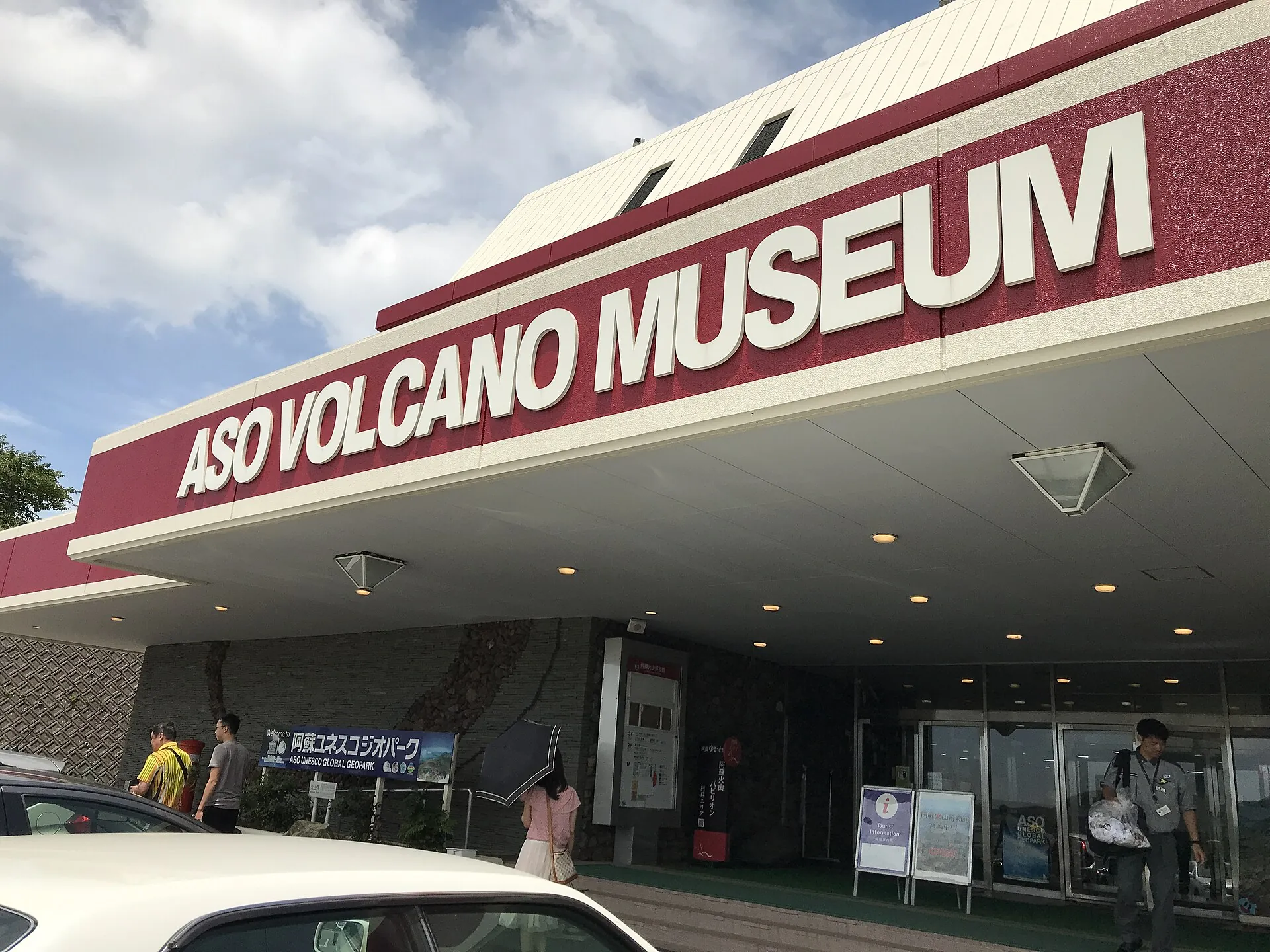 Eingang des Aso Volcano Museum an der Kusasenri mit rotem Schriftzug auf dem Vordach, davor Besucher