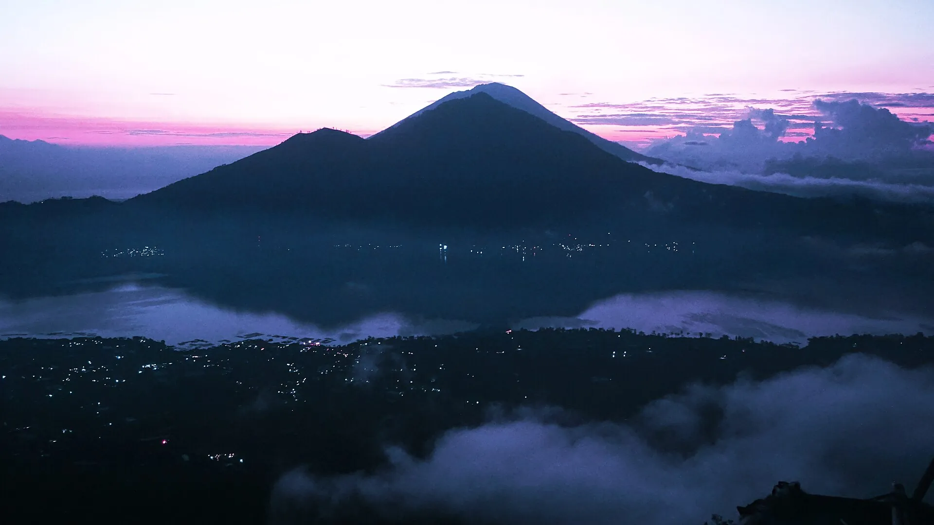 First light over the Batur caldera on Bali: dark mountain silhouettes against a pink sky, with mist banks and village lights below