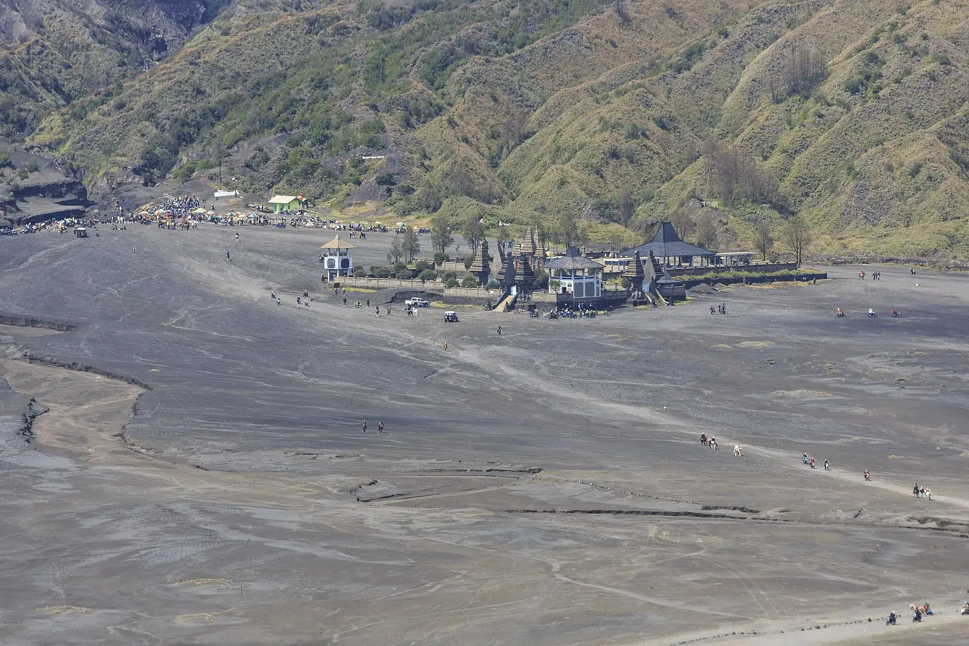 The Hindu temple Pura Luhur Poten in the gray Sea of Sand of the Tengger Caldera, green caldera walls behind it, jeeps and visitors all around