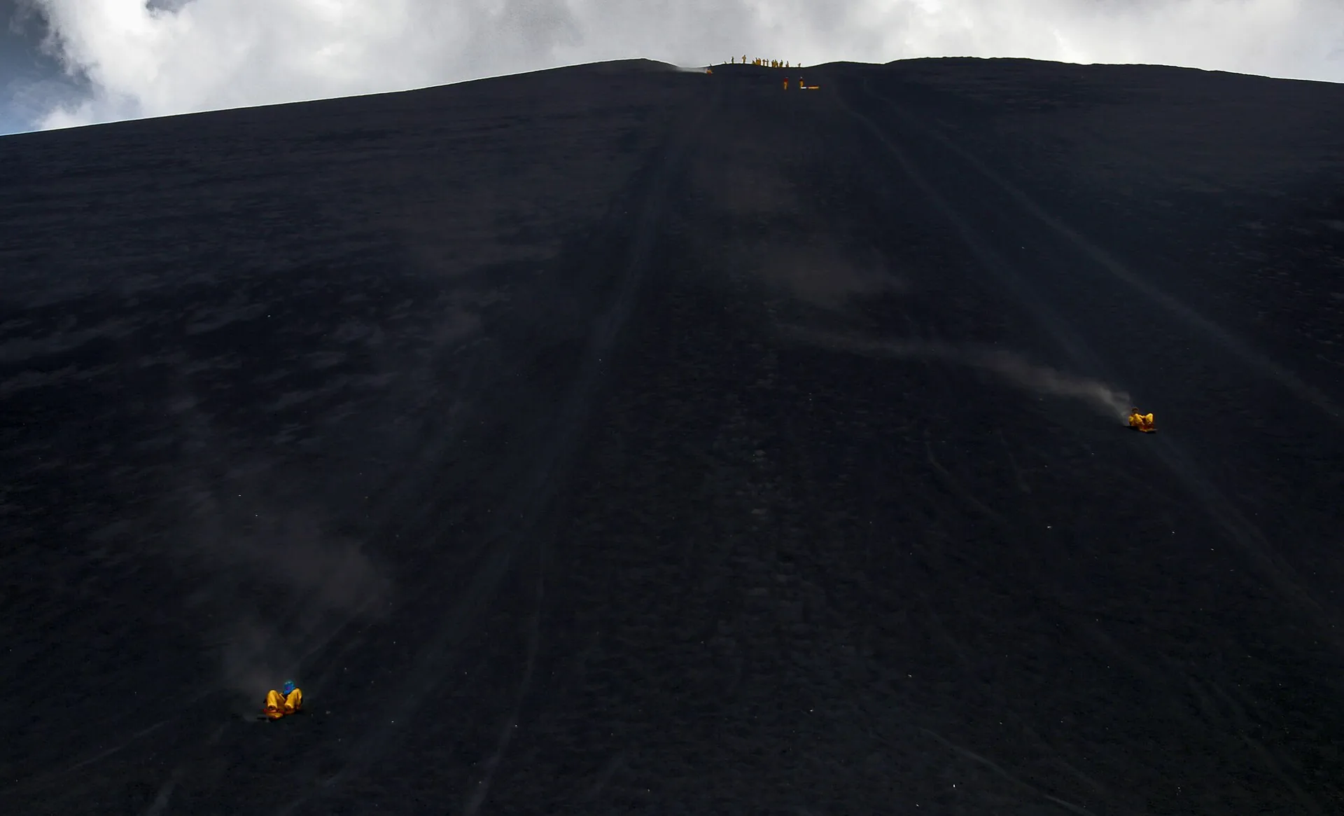Zwei Boarder in orangefarbenen Overalls rutschen die schwarze Aschenflanke des Cerro Negro hinunter