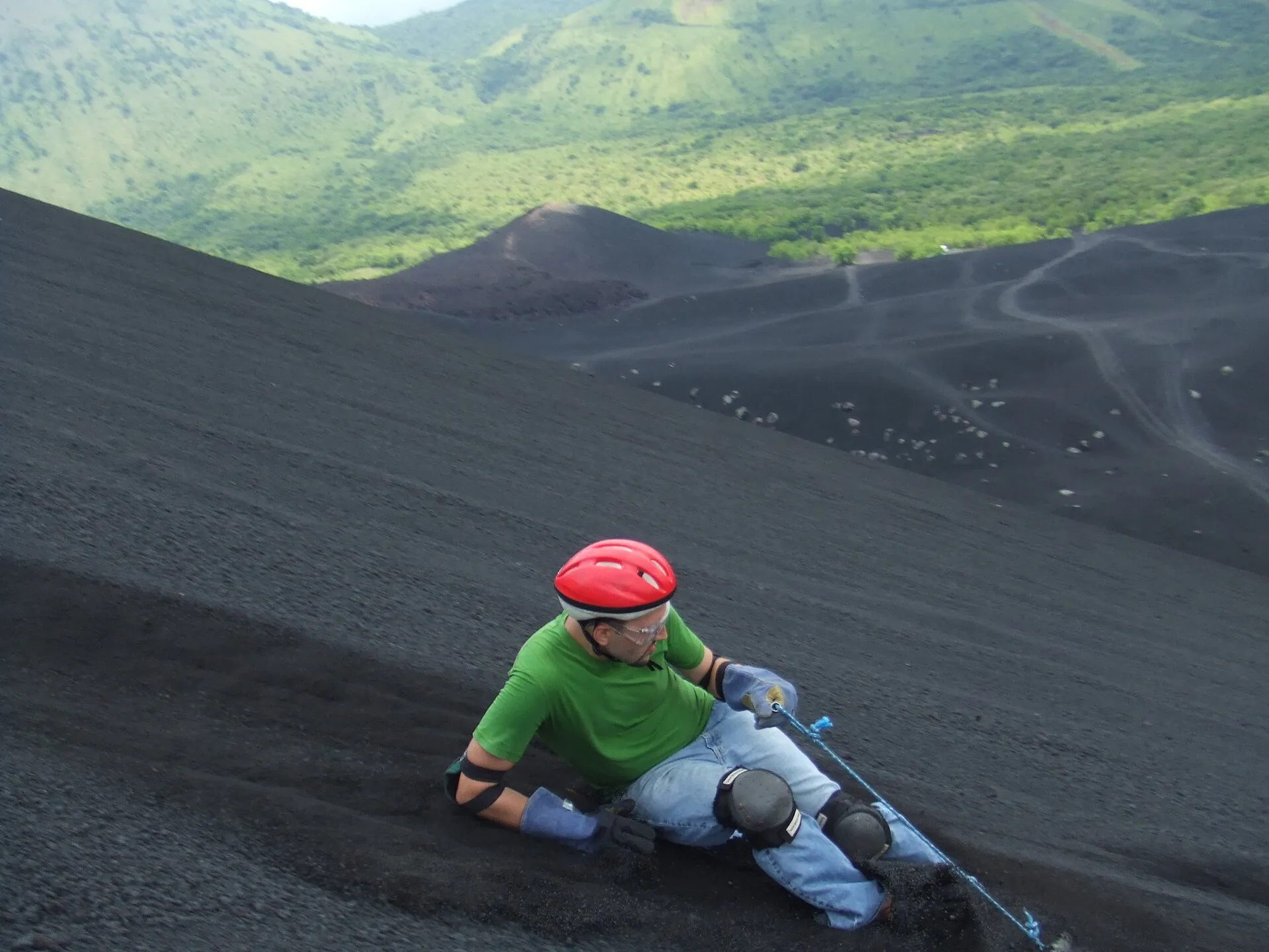 Ein Boarder mit rotem Helm, grünem Shirt und Knieschützern sitzt auf dem Brett und rutscht die Aschenflanke des Cerro Negro hinunter
