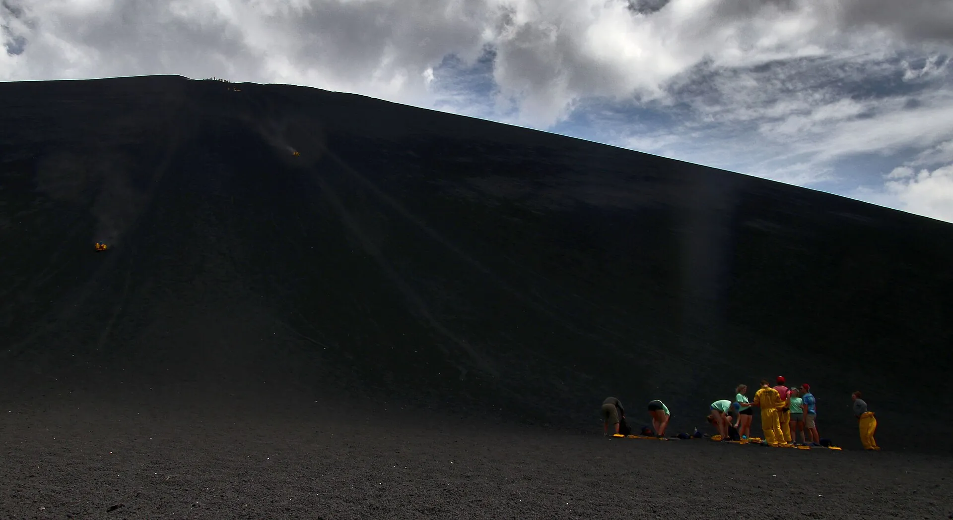 Eine Gruppe in gelben und grünen Overalls sammelt sich am Fuß der schwarzen Rutschflanke des Cerro Negro, darüber Wolken