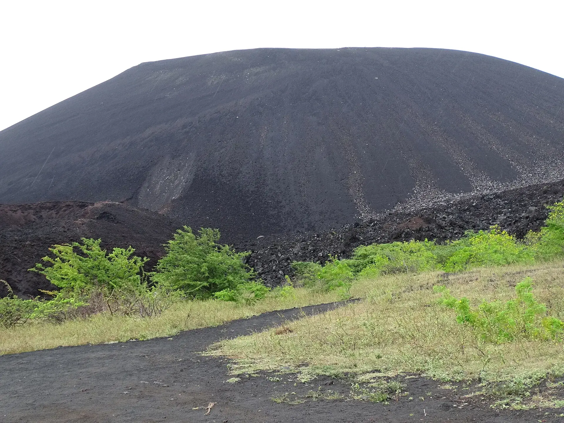 Der pechschwarze Schlackenkegel des Cerro Negro bei León vom Fuß aus, davor grünes Buschwerk auf dunkler Asche
