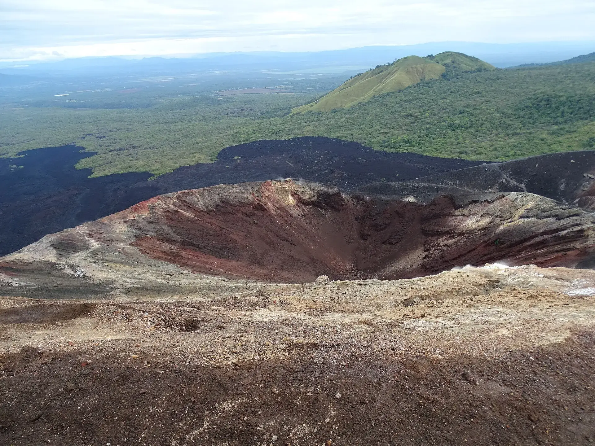 Der Krater des Cerro Negro von oben: rote und schwarze Schlackenwände, dahinter die grüne Ebene und die Berge bei León