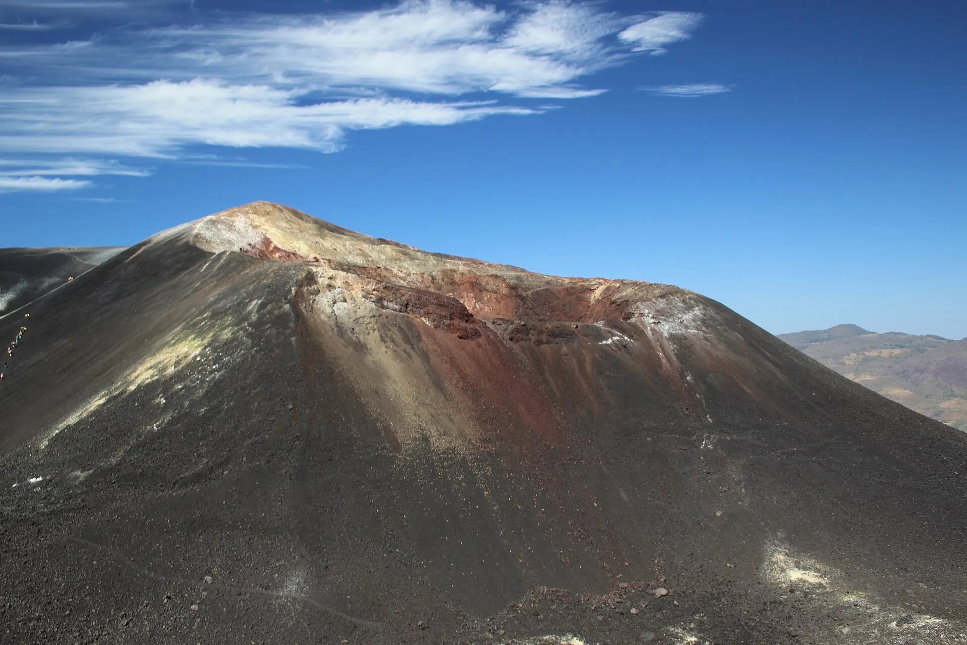 Der Cerro Negro unter tiefblauem Himmel mit Wolken: schwarzer Kegel, Kraterrand rot und weiß von Schwefel