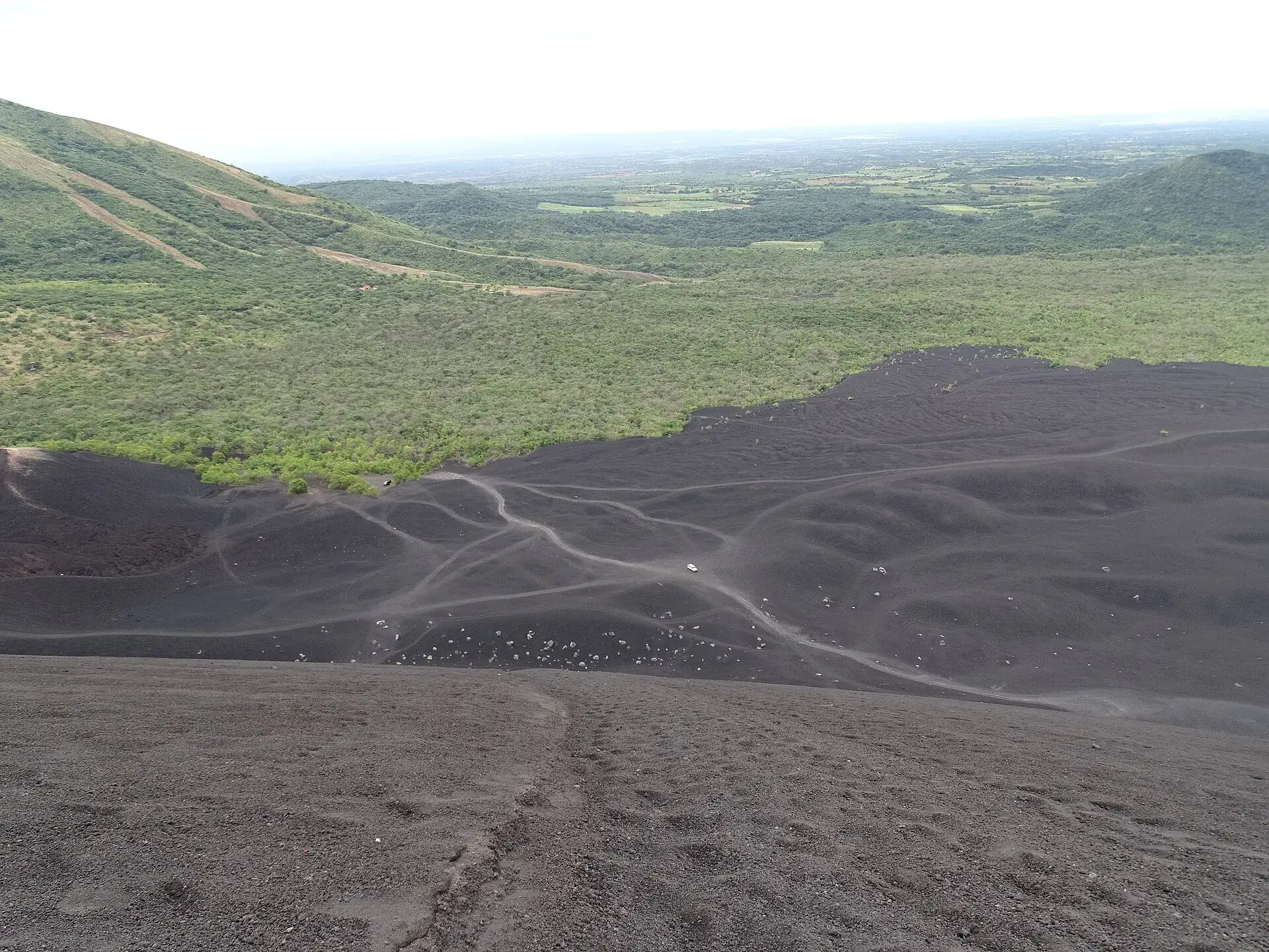 Die Rutschflanke des Cerro Negro von oben: grauschwarze Asche mit den hellen Spuren der Boarder, dahinter grüne Hügel