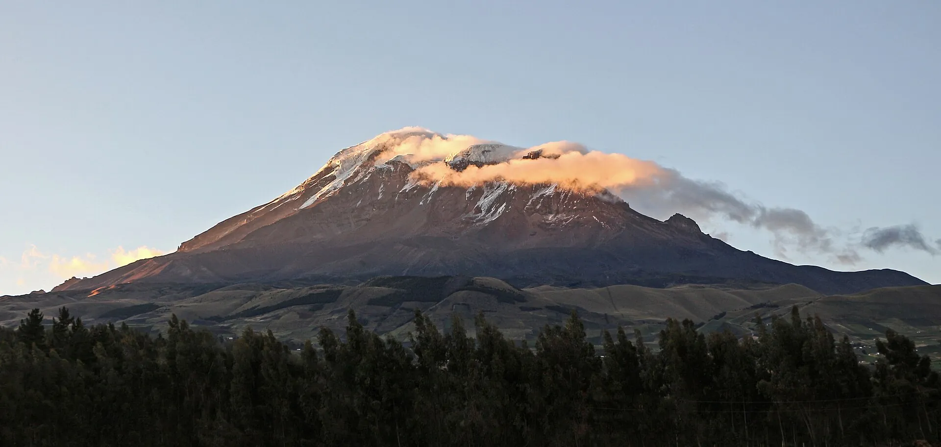 Der Gipfel des Chimborazo im rosa Abendlicht über dunklen Wäldern, der Rest des Berges schon im Schatten
