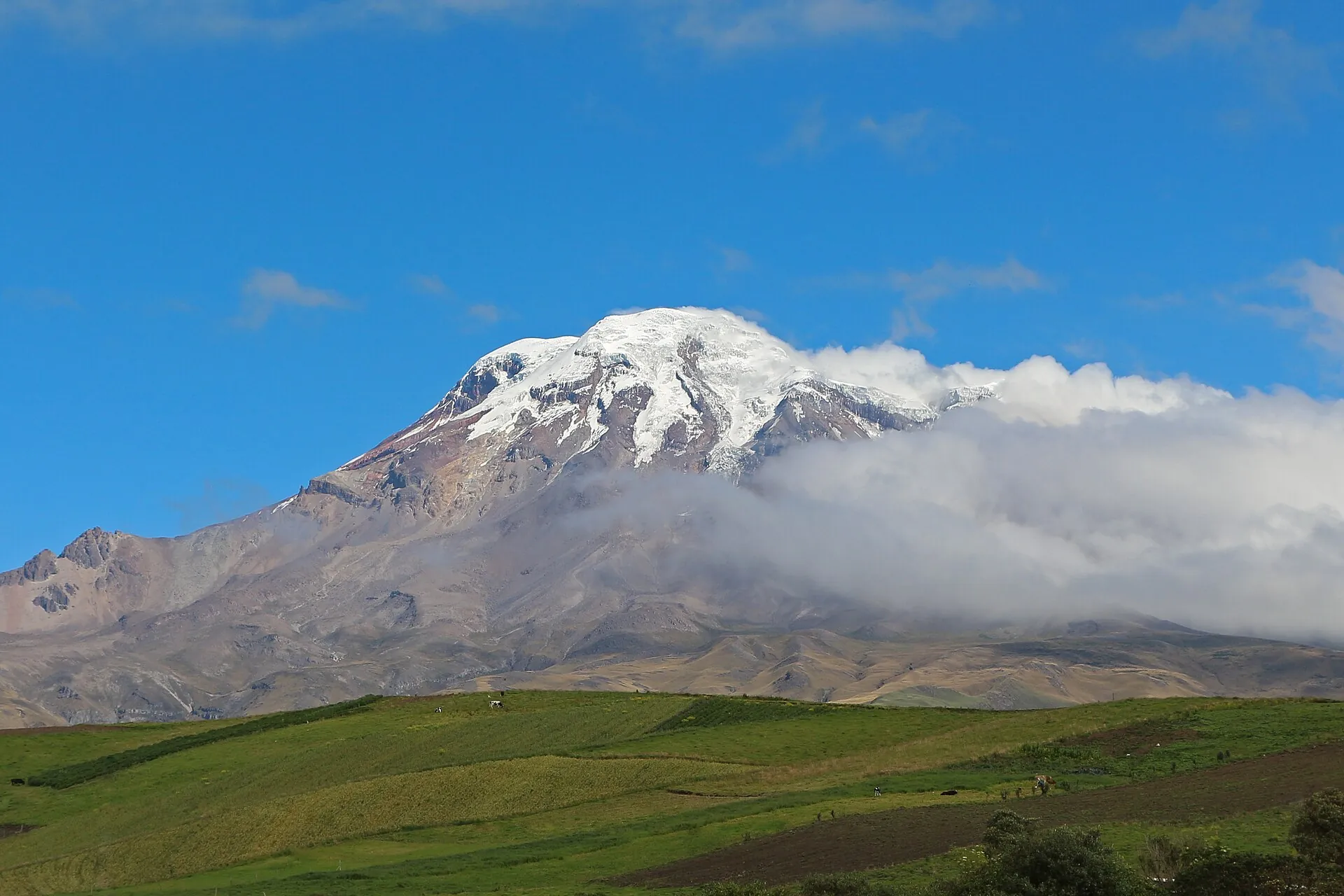 Der vergletscherte Chimborazo hinter grünen Feldern, Wolken hängen an seiner Flanke