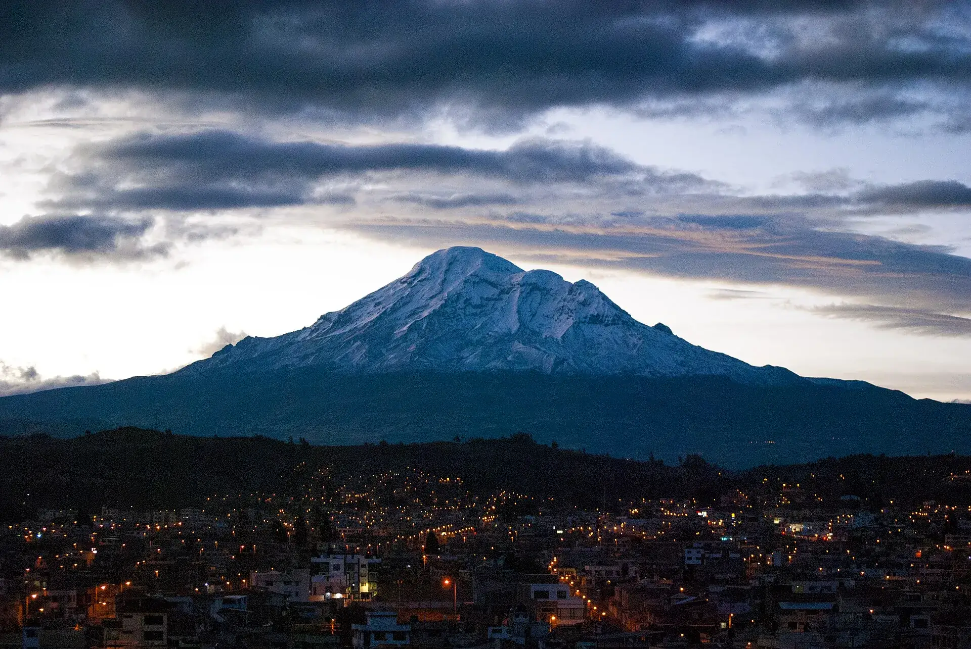 Der Chimborazo in der Abenddämmerung über den Lichtern von Riobamba, dunkle Wolken am Himmel