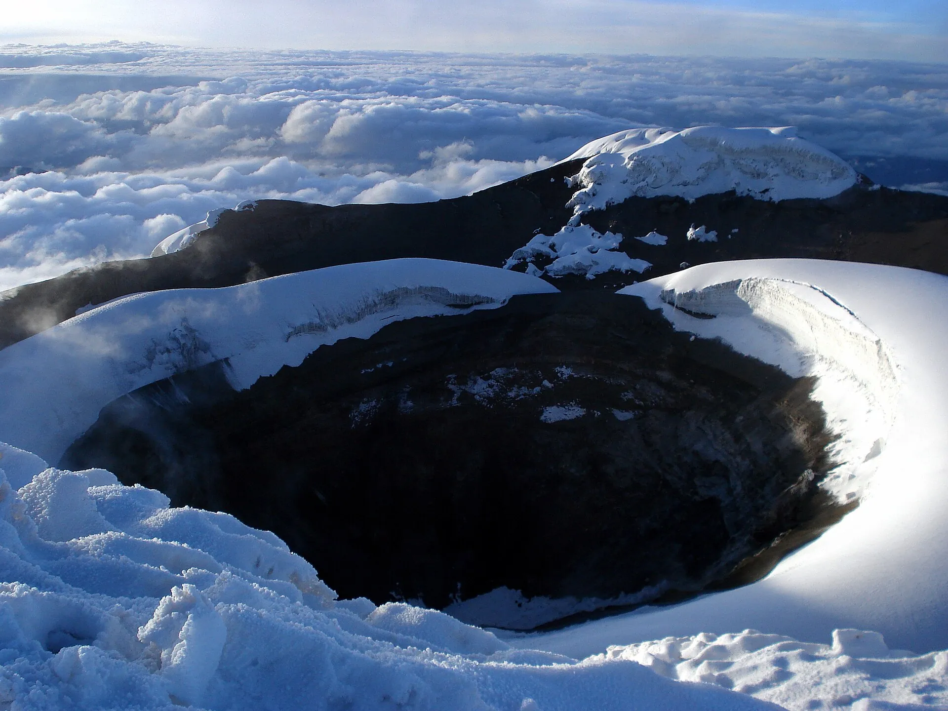 Der Gipfelkrater des Cotopaxi: ein dunkler Schlund mit steilen Eiswänden, ringsum Schnee und Wolken