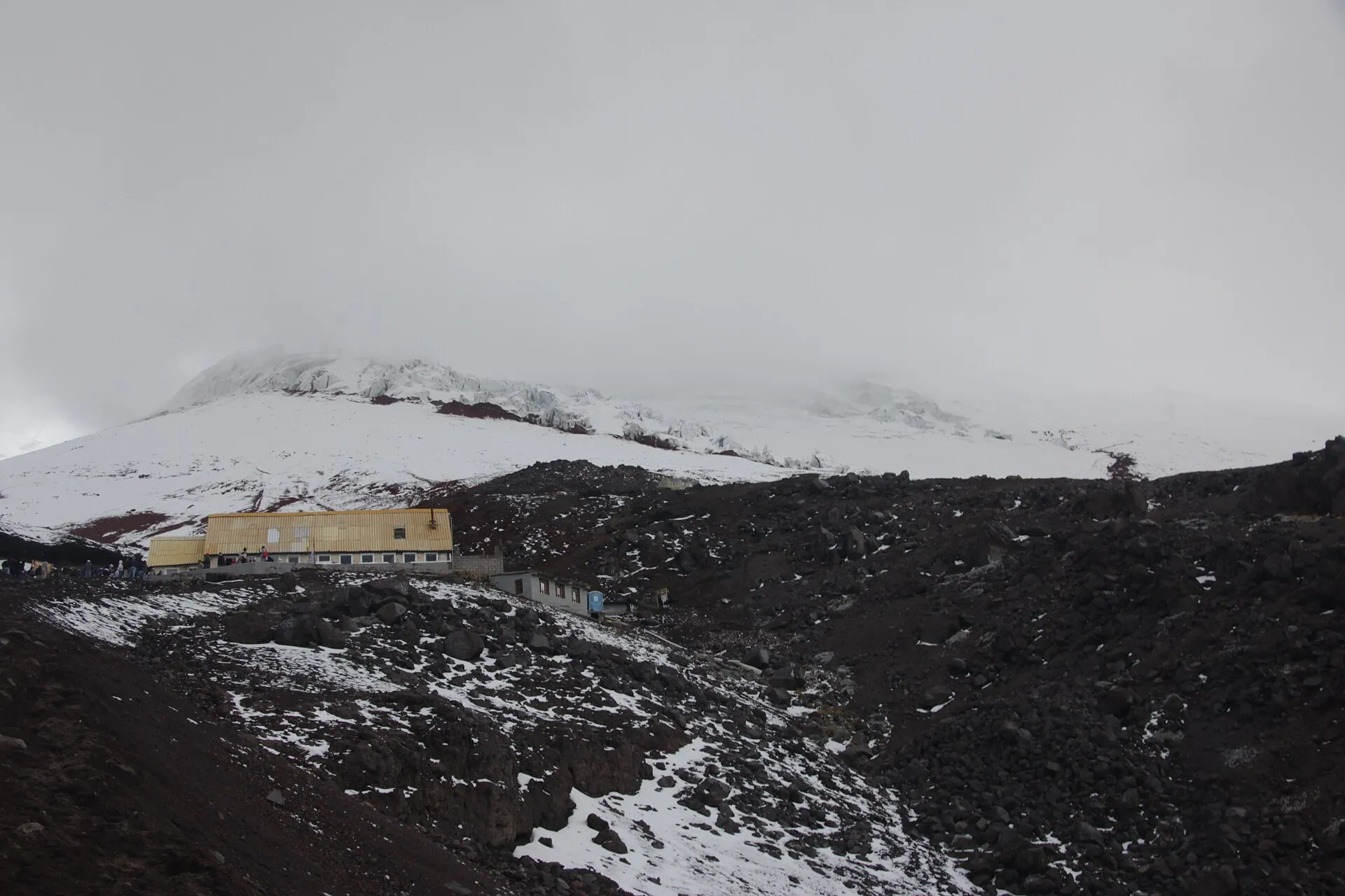 Das Refugio José Rivas als kleines Gebäude auf dem dunklen Aschehang des Cotopaxi, oben Schnee und Wolken