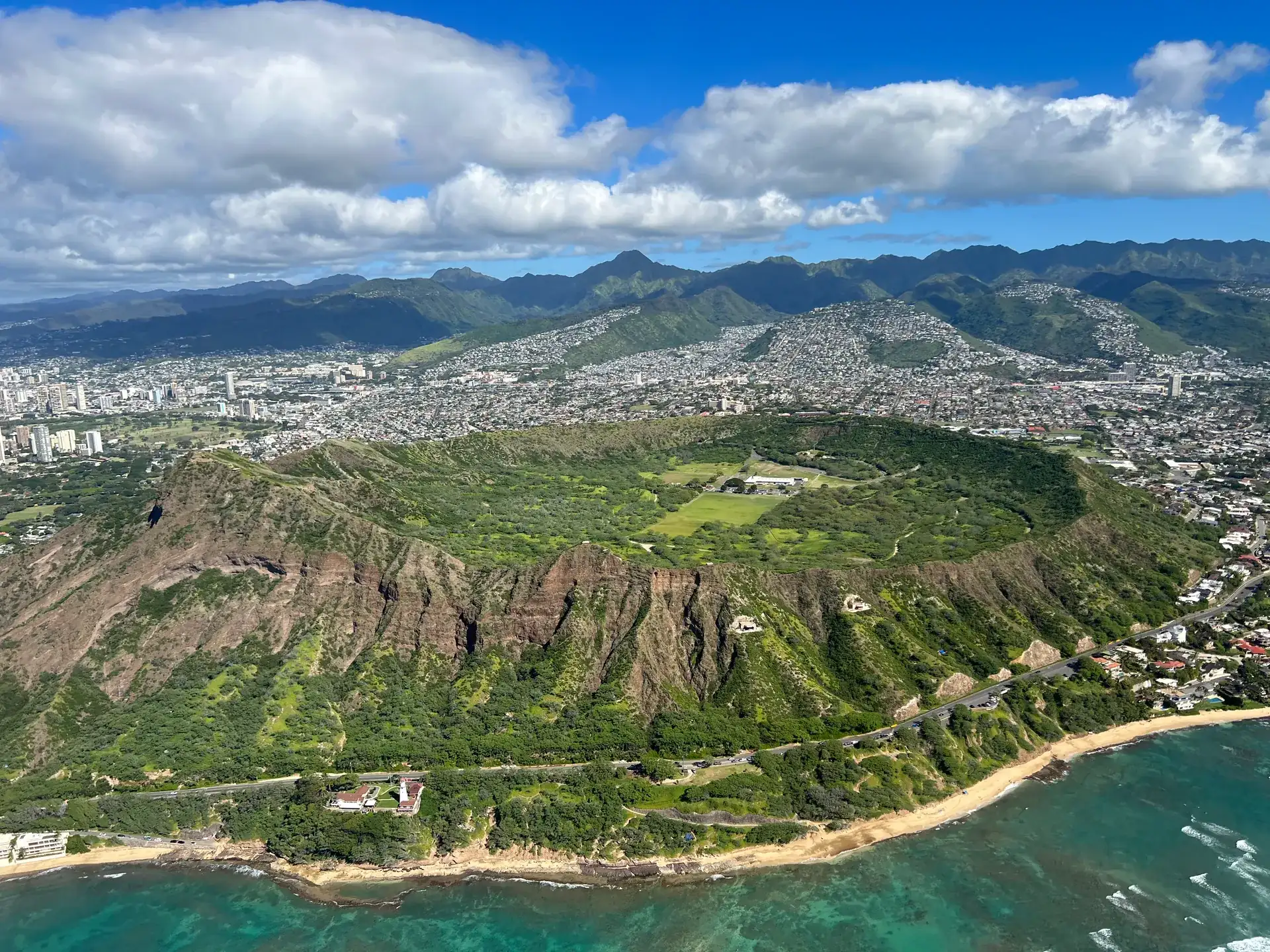 Aerial view of Diamond Head on Oʻahu: a closed, green crater ring right on the coast, with Honolulu and the Koʻolau mountains behind