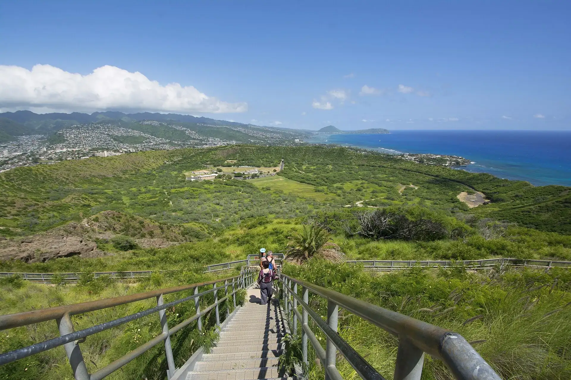 Metal staircase on the Diamond Head Summit Trail with hikers, the green crater floor, the crater rim and the ocean behind