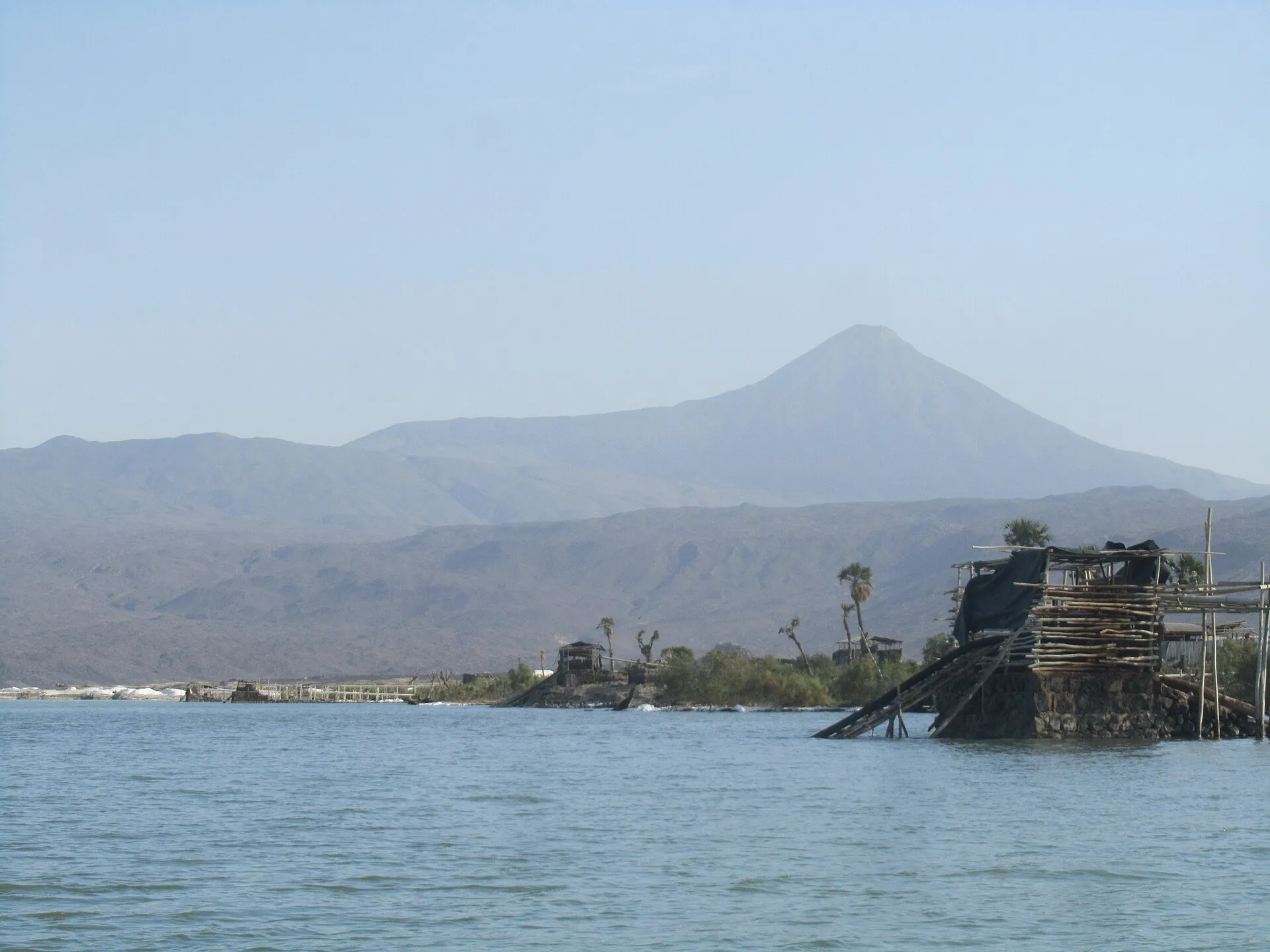 Lake Afdera in Ethiopia with a salt works on the shore and the cone of Afdera volcano in the haze behind