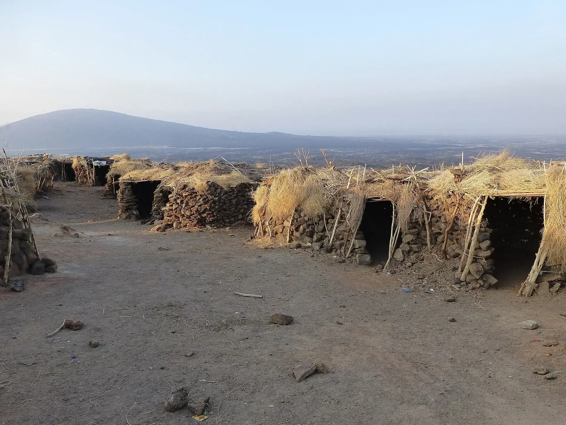 Several thatched stone huts on the crater rim of Erta Ale in morning light, the flat Danakil plain beyond