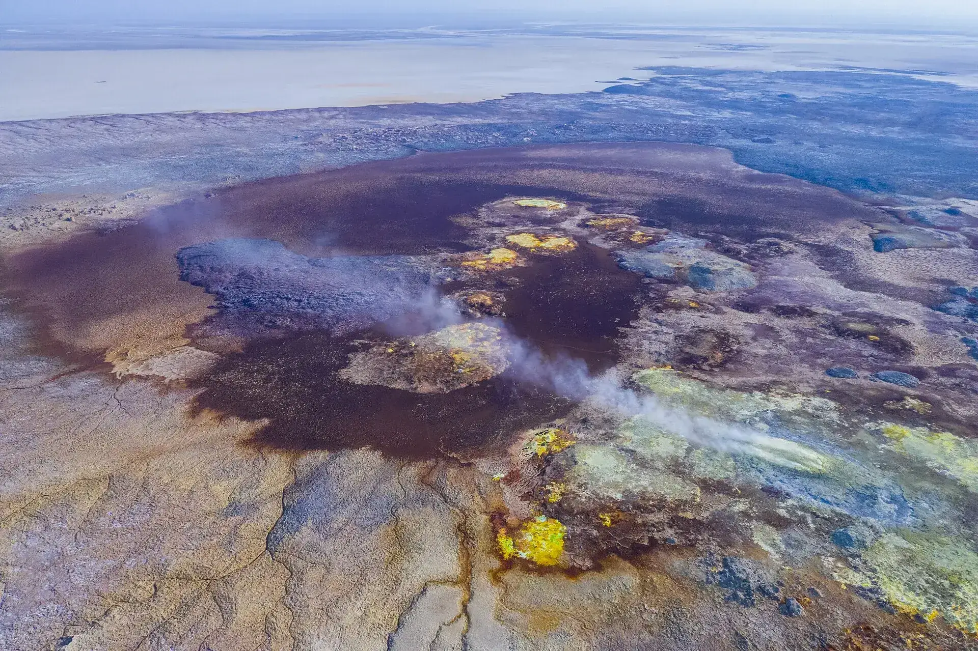 Aerial view of the Dallol hydrothermal field: yellow, green and rust-red salt terraces around a dark crater, steam above the springs