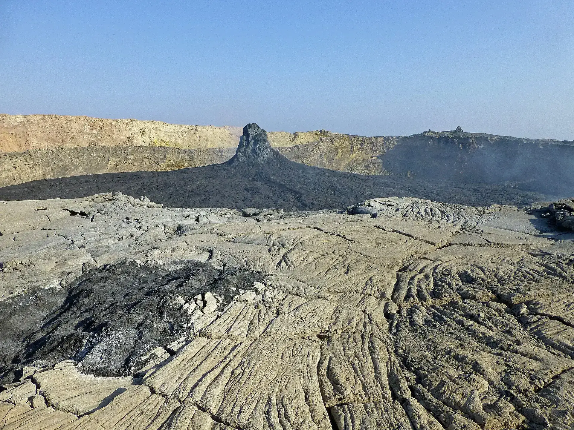 The crater floor of Erta Ale: a dark hornito rises from frozen lava, steam drifting from the collapsed north crater on the right