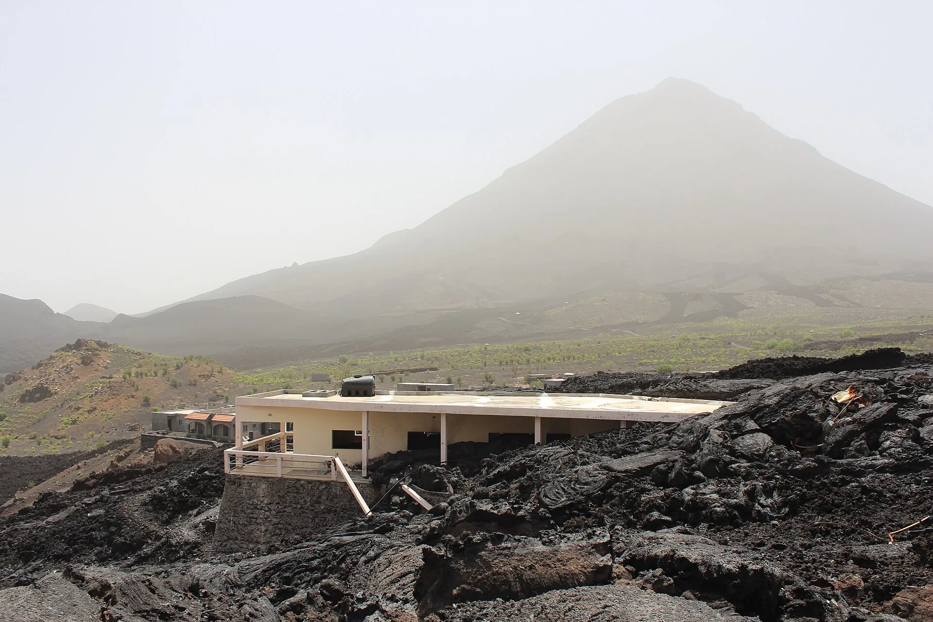 Der Kegel des Pico do Fogo im Dunst der bruma seca über den Ruinen eines von Lava umschlossenen Hauses in Chã das Caldeiras