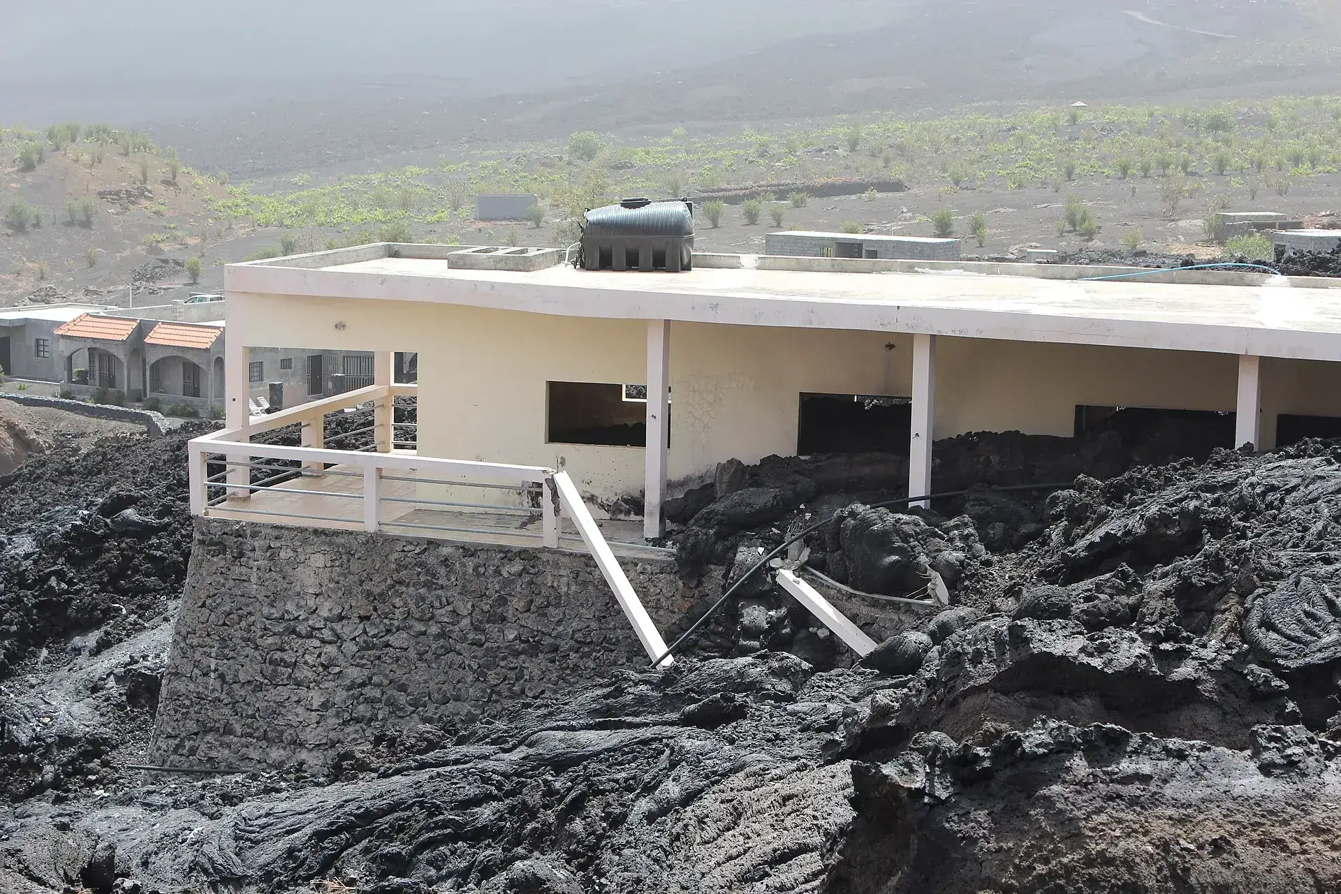 Ein Haus in Portela auf Fogo, dessen Erdgeschoss von der schwarzen Lava des Ausbruchs 2014/15 umschlossen ist, dahinter Reben und die Caldera