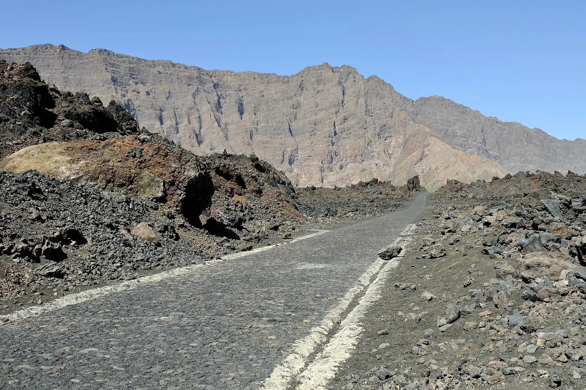 Die gepflasterte Straße in die Caldera Chã das Caldeiras auf Fogo, links schwarze Lava, dahinter die Steilwand der Bordeira