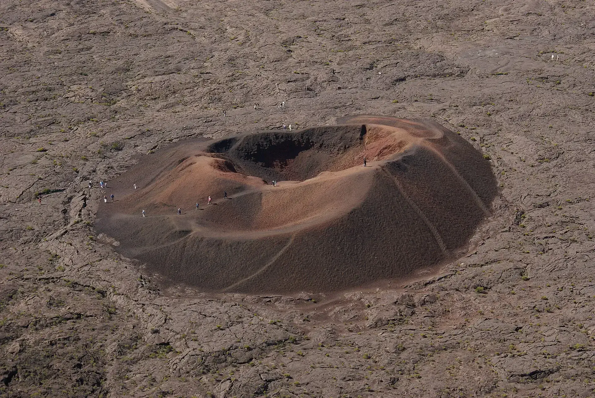 Formica Léo, a small red double cinder cone in the middle of the gray lava field of the Enclos Fouqué, tiny hikers at its edge