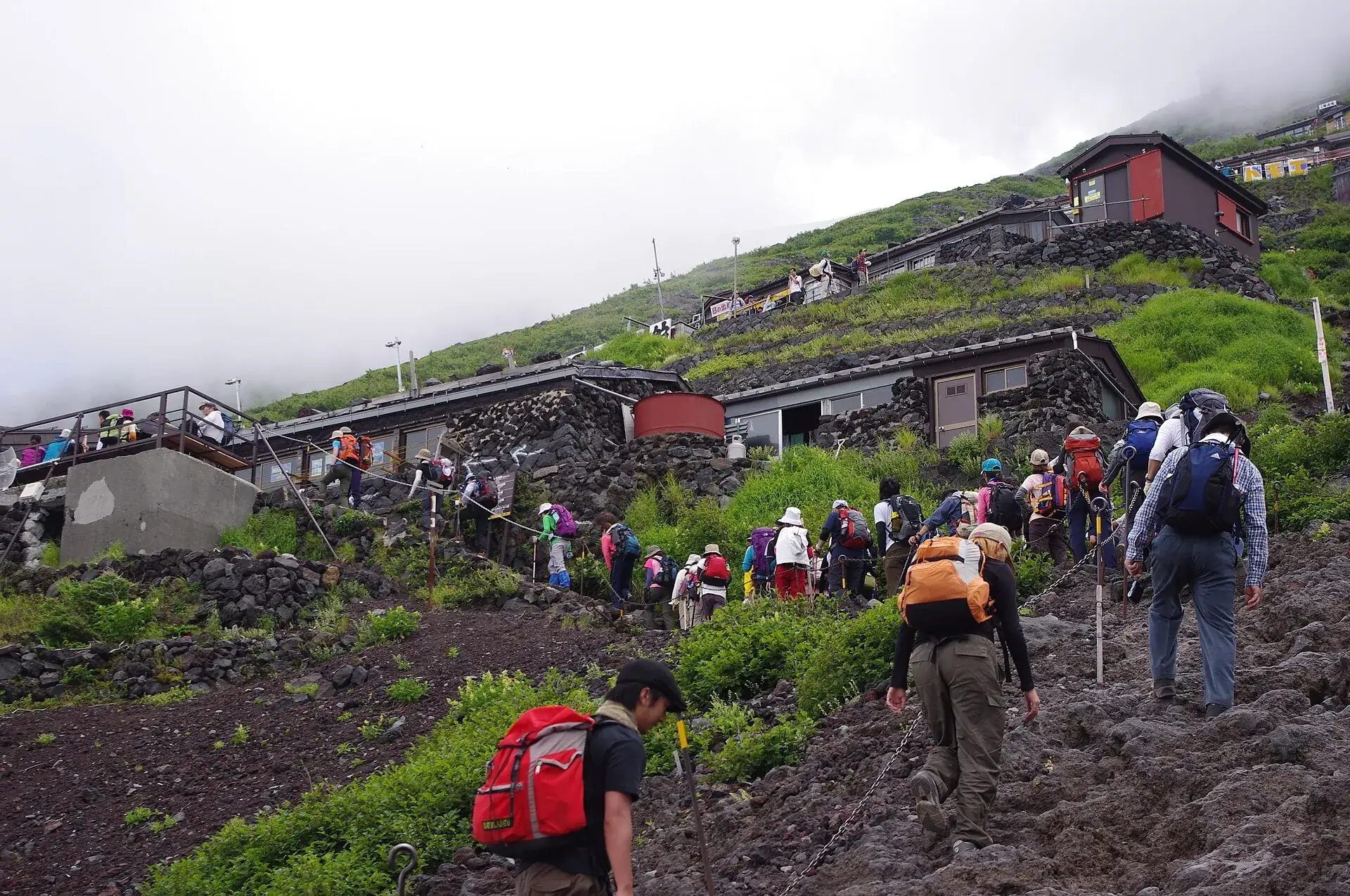 Climbers on the Yoshida Trail on Mount Fuji: lava gravel, green slopes and mountain huts disappearing into cloud