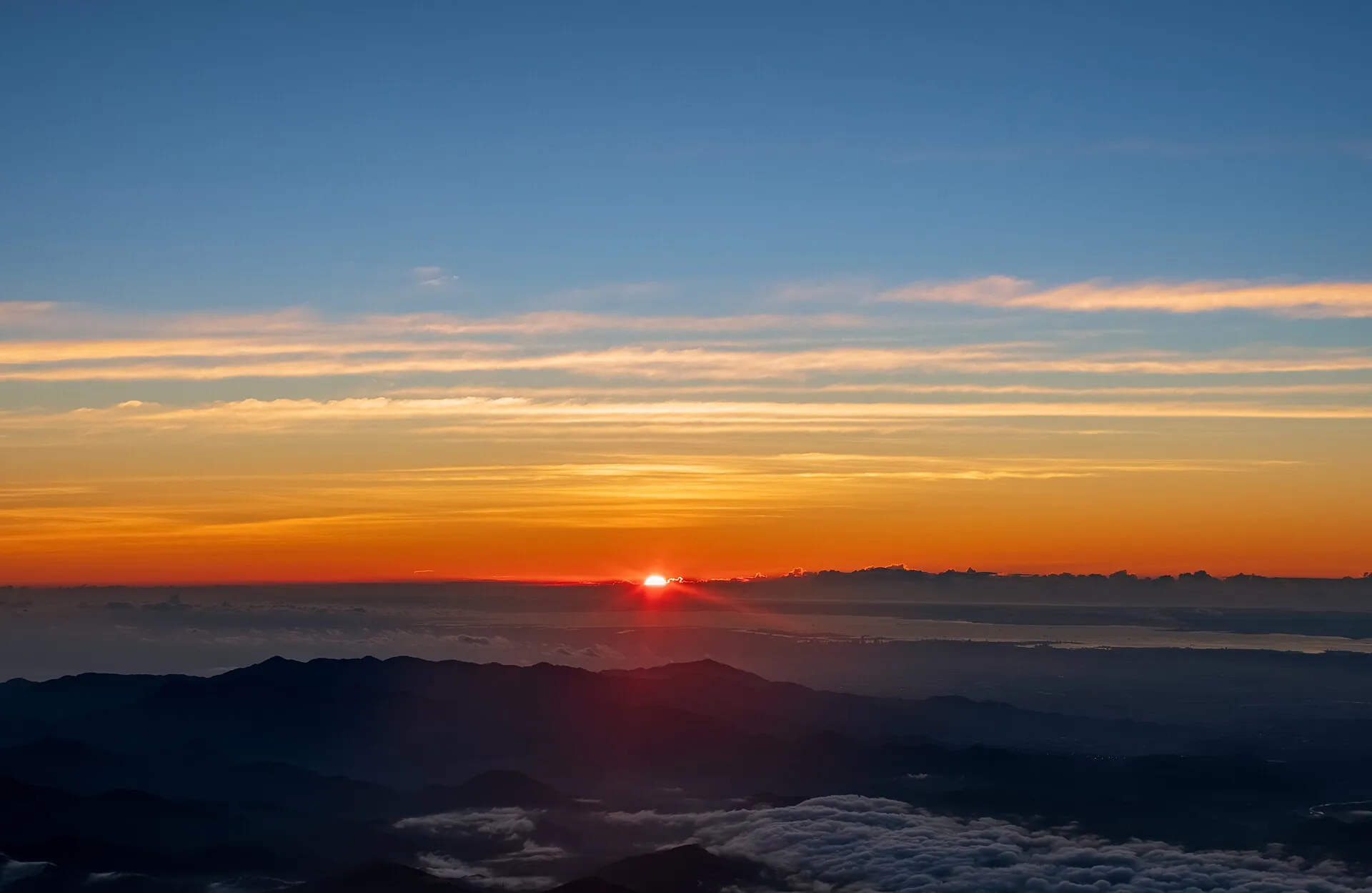 Sunrise from the summit of Mount Fuji: the sun rising red above the horizon, mountain ranges and clouds below