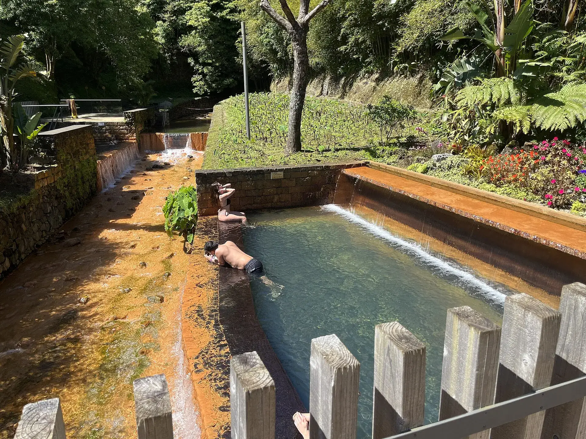 Ein Thermalbecken der Poça da Dona Beija in Furnas: eisenbraunes Wasser zwischen Steinmauern, Holzpfosten am Rand, Gäste im Wasser
