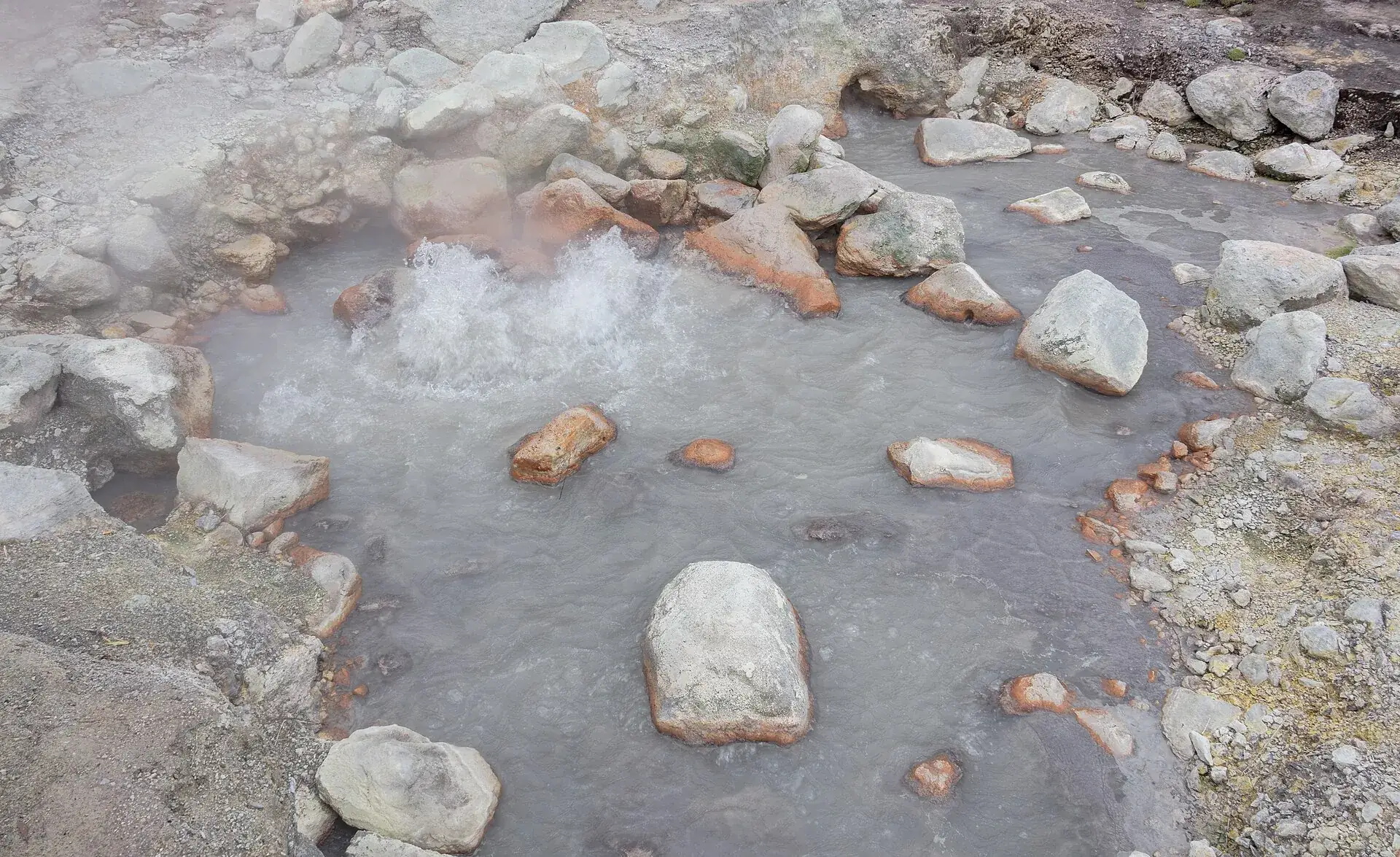 Eine kochende Quelle in Furnas: graues Wasser blubbert zwischen rot verkrusteten Steinen