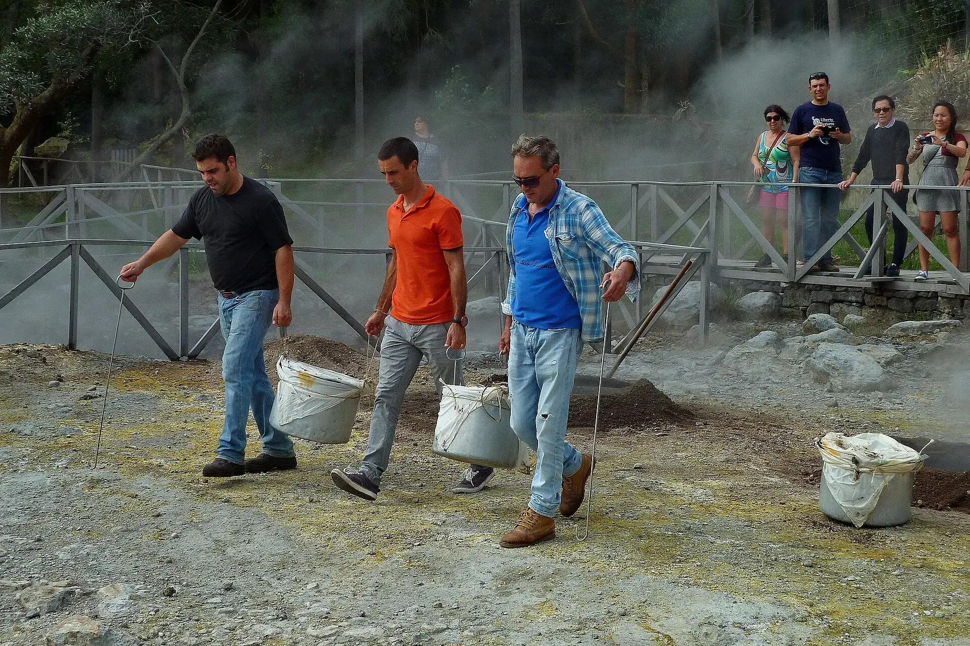 Zwei Männer tragen einen dampfenden Cozido-Topf an einer Stange aus der Kochgruben-Zone an der Lagoa das Furnas, Zuschauer hinter dem Geländer