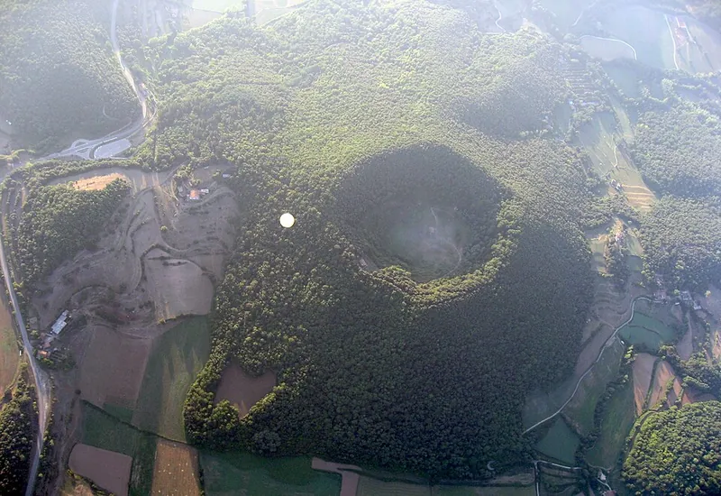 Blick aus einem Heißluftballon auf den bewaldeten Krater des Santa Margarida in der Garrotxa, ein zweiter Ballon schwebt darunter