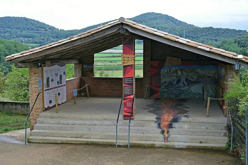 Der offene Holzpavillon des Infozentrums Can Passavent am Croscat mit Schautafeln, dahinter bewaldete Hügel der Garrotxa
