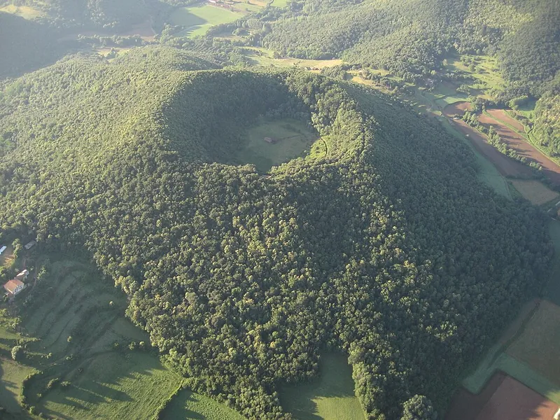 Der kreisrunde, bewaldete Krater des Vulkans Santa Margarida in der Garrotxa aus der Luft, in der Mitte die Wiese mit der Kapelle