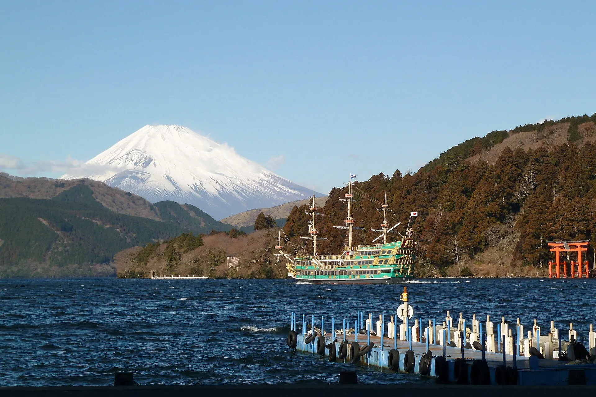 A sightseeing ship styled like a sailing ship on Lake Ashi in Hakone, snow-covered Mount Fuji behind it and a red torii gate on the shore