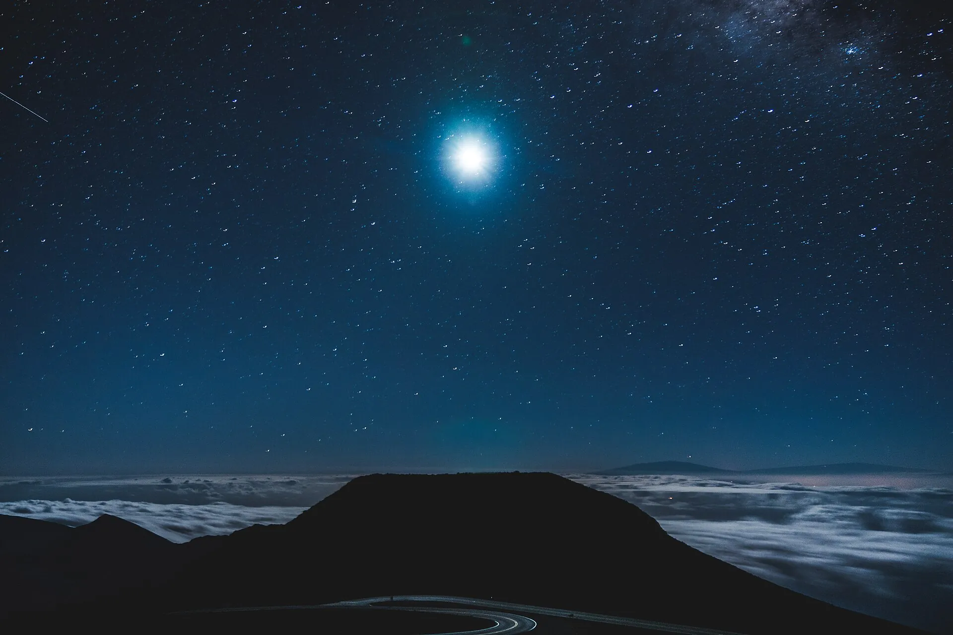 Night at the summit of Haleakalā on Maui: a bright moon and stars above a sea of clouds, with the dark outline of a cone and the summit road in front