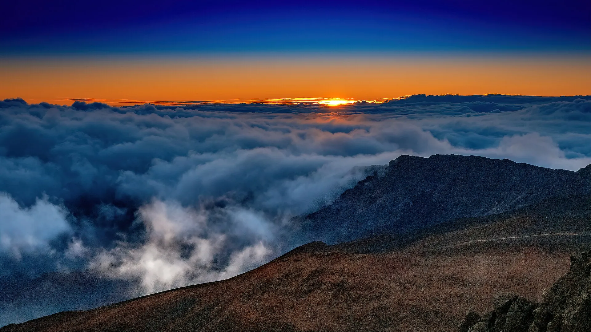 Sunrise at Haleakalā: an orange horizon above a solid layer of clouds, with clouds spilling over the crater rim into the basin
