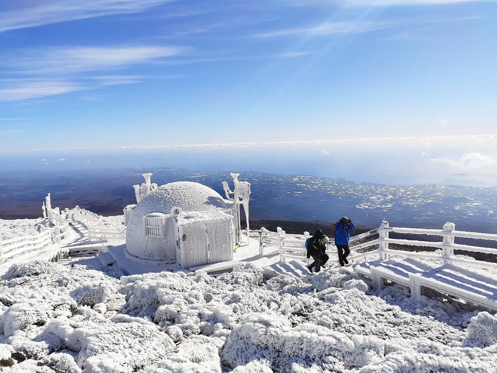 Iced-over summit shelter and railing on Hallasan in winter, hikers in the hoarfrost, the coast of Jeju behind