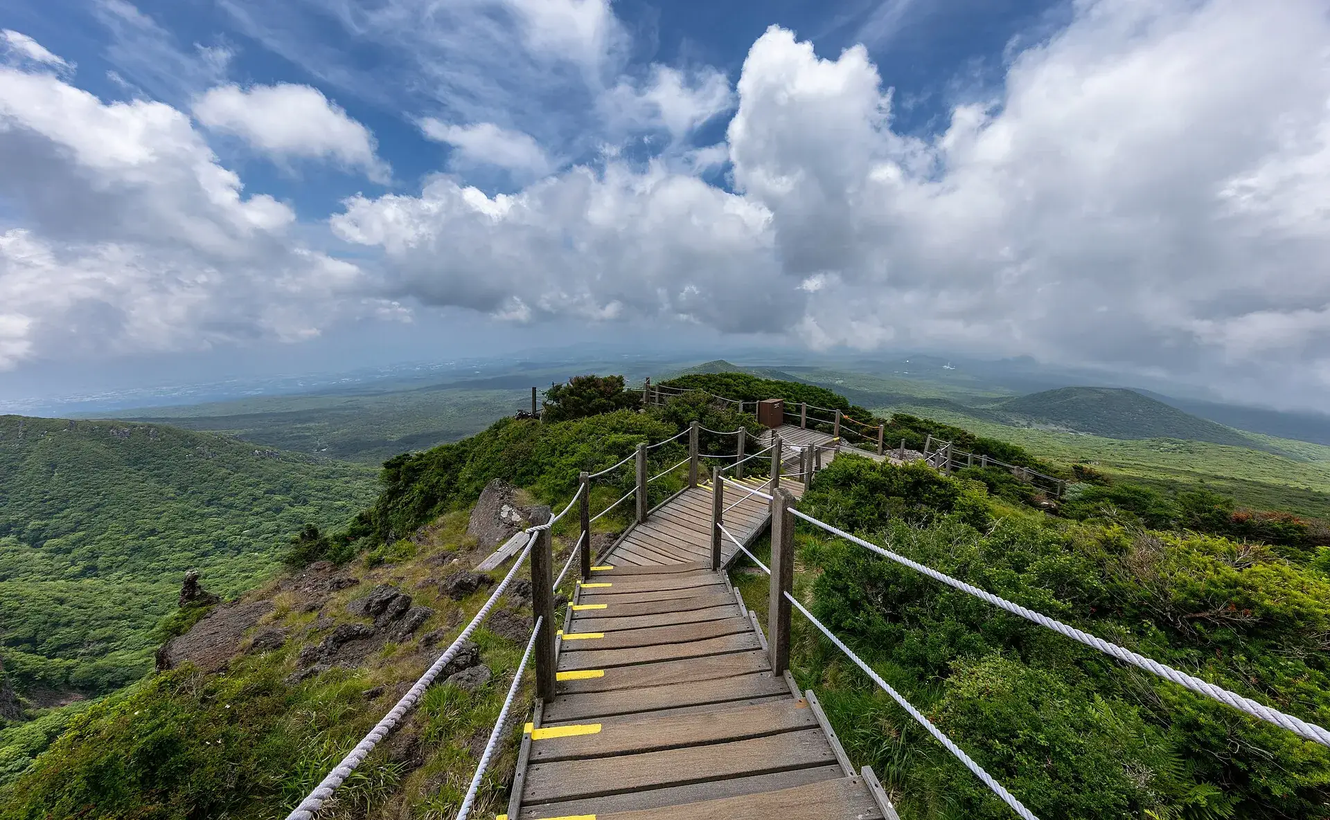 Boardwalk with a rope railing on the Yeongsil Trail, with the mountains of Hallasan National Park and clouds behind