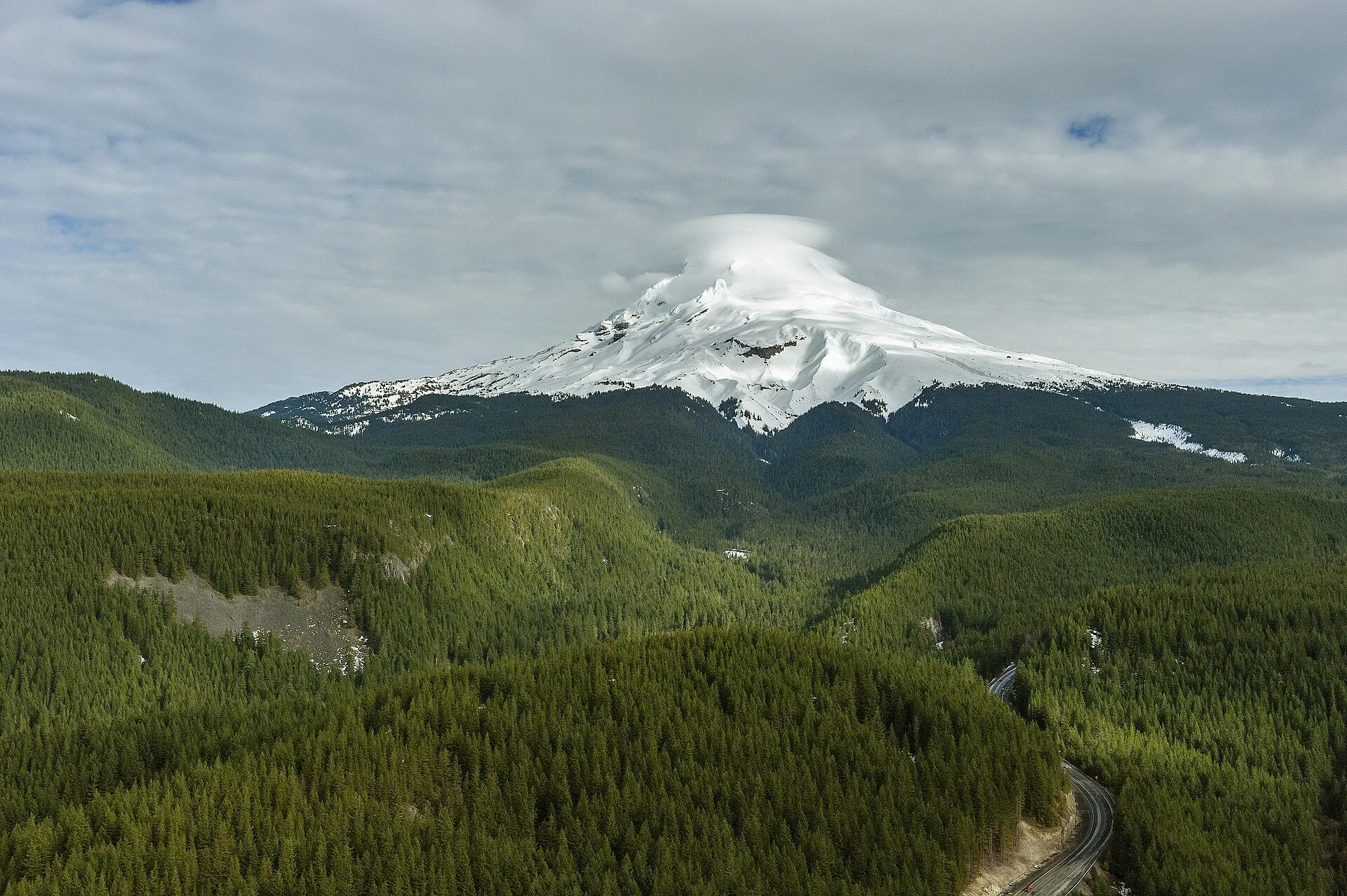 Mount Hood im Winter mit einer linsenförmigen Wolke über dem Gipfel, davor bewaldete Hügel und die Serpentinen der US 26