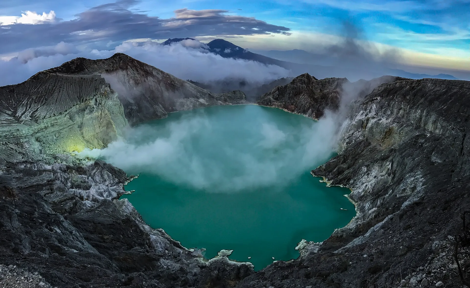 The turquoise acid lake of Kawah Ijen in the morning light, steam drifting over the water, more volcanic cones in the background