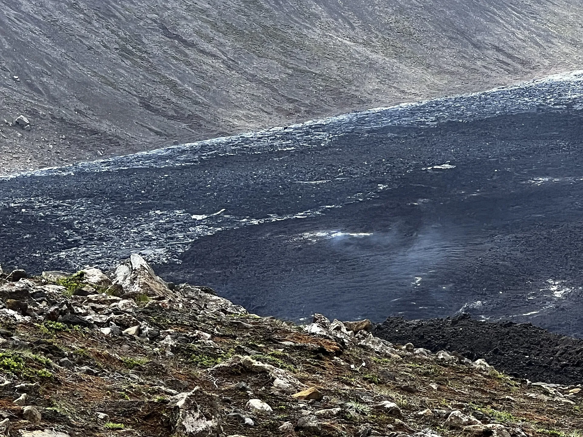 A fresh, glossy black lava field fills a valley near Geldingadalir, still steaming at the edge, a grey slope behind