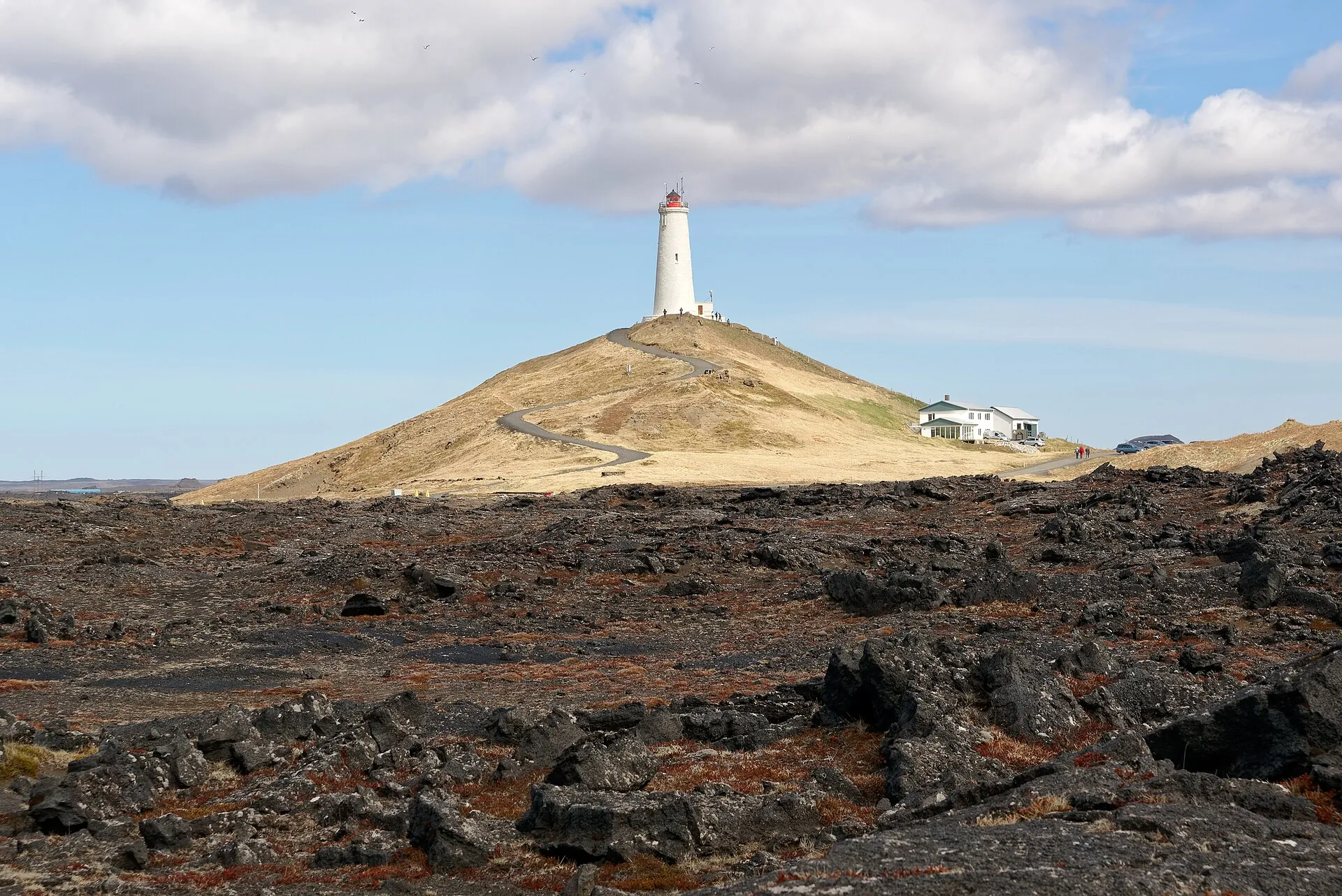 The white Reykjanesviti lighthouse on a grassy hill above an old lava field, clouds in the sky