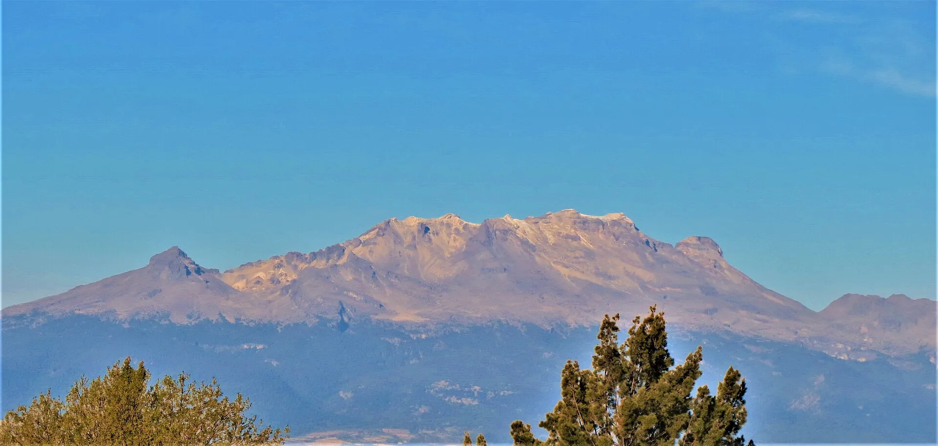 Der Iztaccíhuatl von Osten, von Puebla aus: lange, schneebedeckte Bergsilhouette mit mehreren Gipfeln über einem Baum