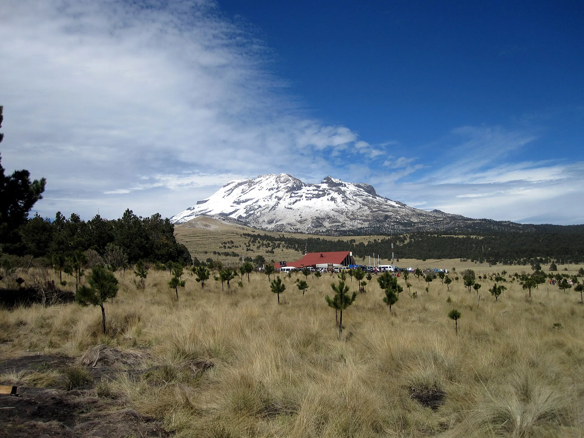 Der schneebedeckte Iztaccíhuatl über einer Hochlandwiese mit jungen Kiefern und einem Gebäude mit rotem Dach am Paso de Cortés